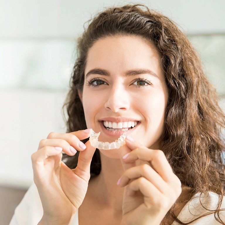 Young woman holding clear dental aligners, smiling in bright indoor setting.