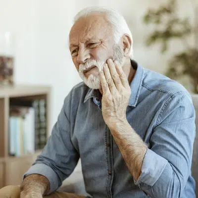 Senior man touching his cheek in pain, sitting indoors in casual blue shirt.