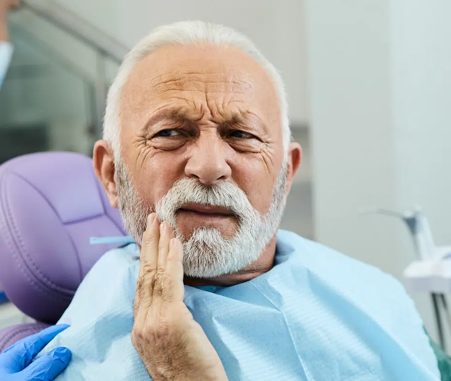 Senior man with hand to cheek experiencing tooth pain in dental office chair.